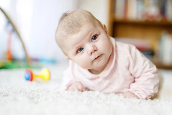 Adorable newborn baby girl lying on belly in white sunny bedroom or nursery. Textile and bedding for children. Cute girl with blue eyes at home playing with colorful rattle toys. — Stock Photo, Image