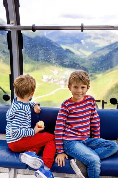 Two little boys sitting inside of cabin of cable car and looking on mountains landscape. Family on hiking vacations in Austria. Siblings having fun with exploring glacier and forest from window. — Stock Photo, Image