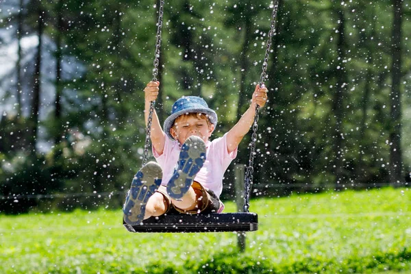 Garoto engraçado garoto se divertindo com balanço de corrente no parque infantil ao ar livre enquanto está molhado salpicado com água. criança balançando no dia de verão. Lazer ativo com crianças. Menino chorão feliz com gotas de chuva no rosto. — Fotografia de Stock