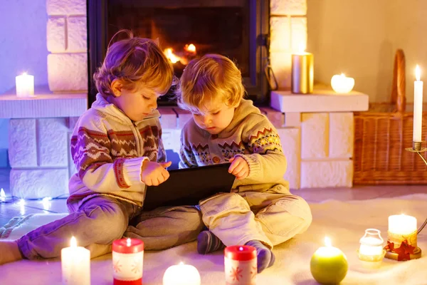 Δύο μικρά παιδιά κάθονται δίπλα σε ένα τζάκι στο σπίτι τα Χριστούγεννα. Ευτυχισμένα χαριτωμένα αξιολάτρευτα μικρά αγόρια, ξανθά δίδυμα που παίζουν με το νέο δώρο tablet. Οικογένεια γιορτάζει Χριστούγεννα διακοπές. — Φωτογραφία Αρχείου