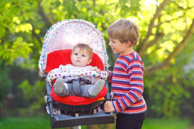 Küçük sarışın çocuk çocuk kardeşim ile oynamak. Mutlu kardeşler Bahçe. Çocuk arabası ya da bebek arabası oturan kız bebek. Kardeşim ve yaz gününde gidiş birlikte bir yürüyüşe şirin toddler açık havada