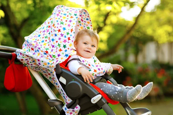 Leuke gezonde kleine mooie baby meisje zitten in de kinderwagen of kinderwagen en wachten op moeder. Gelukkig lachend kind met blauwe ogen. Met groene boomachtergrond. Kleine dochter gaat wandelen met familie — Stockfoto