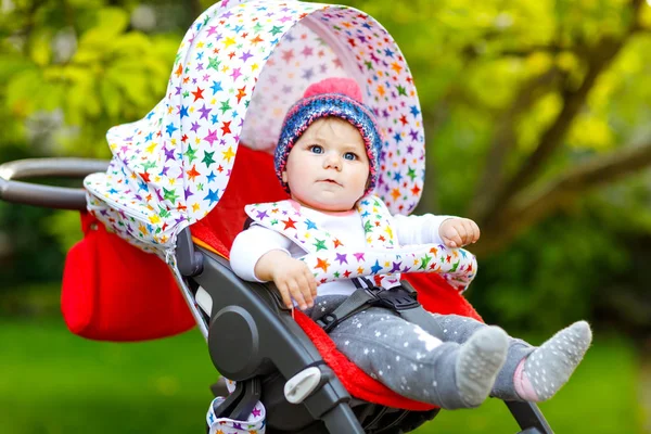Cute healthy little beautiful baby girl with blue warm hat sitting in the pram or stroller and waiting for mom. Happy smiling child with blue eyes. baby daughter going for a walk with family — Stock Photo, Image