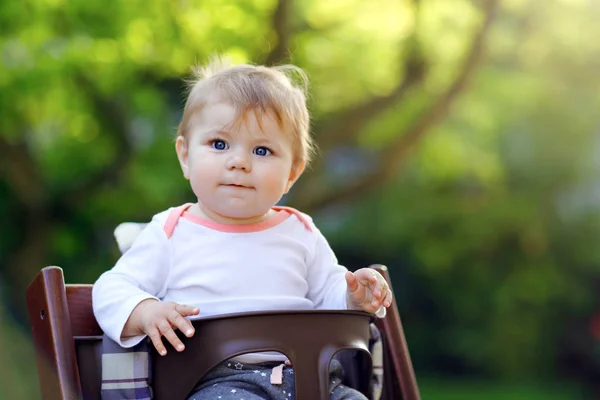 Söt bedårande baby flicka sitter i barnstol utomhus. Beatuiful barn 6 månader i hem trädgård, spelar på varm solig dag. Friskt barn ler och skrattar. — Stockfoto