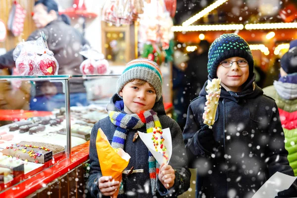 Dois garotinhos, irmãos bonitos comendo bananas cobertas de chocolate, marshmellows e polvilhas coloridas perto de doce stand com pão de gengibre e nozes. Crianças felizes no mercado de Natal na Alemanha . — Fotografia de Stock