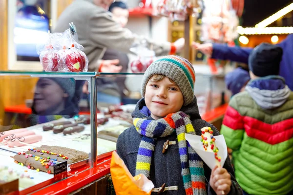 Menino, criança fofa comendo bananas cobertas de chocolate, marshmellows e polvilhas coloridas perto de doce stand com pão de gengibre e nozes. Menino feliz no mercado de Natal na Alemanha . — Fotografia de Stock