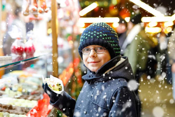 Menino, criança fofa comendo bananas cobertas de chocolate, marshmellows e polvilhas coloridas perto de doce stand com pão de gengibre e nozes. Menino feliz no mercado de Natal na Alemanha . — Fotografia de Stock