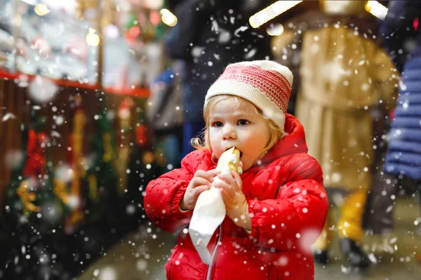 Little baby girl, cute child eating bananas covered with chocolate, marshmellows and colorful sprinkles near sweet stand with gingerbread and nuts. Happy toddler on Christmas market in Germany. — Stock Photo, Image