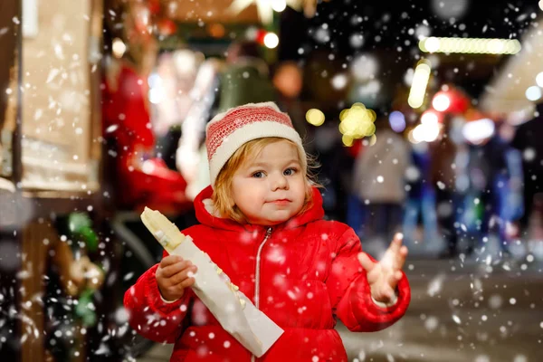 Kleines Mädchen, süßes Kind, das Bananen mit Schokolade, Marshmellows und bunte Streusel in der Nähe von Süßwarenstand mit Lebkuchen und Nüssen isst. Glückliches Kleinkind auf dem Weihnachtsmarkt in Deutschland. — Stockfoto