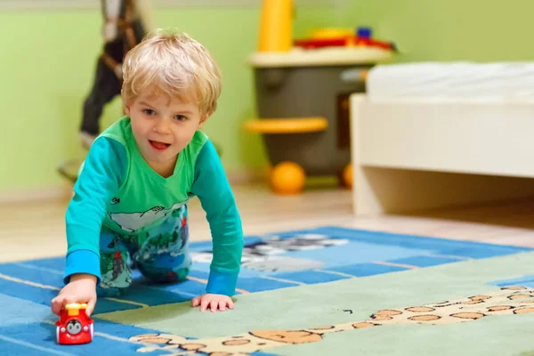 Feliz niño rubio divertido jugando con un montón de coches de juguete en el interior. Niño con camisa de colores y divertirse en la guardería. Niño en ropa de dormir jugando en la habitación en casa . — Foto de Stock