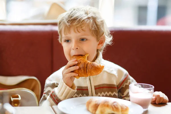 Cute healthy kid boy eating croissant and drinking strawberry milkshake in cafe. Happy child having breakfast with parents or at nursery. Vegetables, eggs as healthy food for children. — Stock Photo, Image