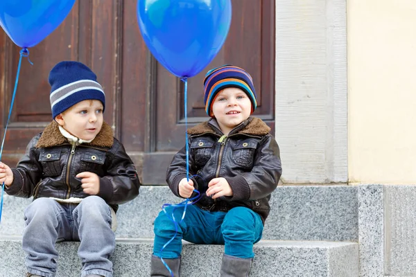 İki küçük çocuklar açık havada mavi hava balonları ile oynayan çocuk. Mutlu ikizler ve gülümseyen gülen ve birlikte yürümeye başlayan kardeşler. Sıcak giysiler içinde soğuk günde dışında sağlıklı çocuk. — Stok fotoğraf