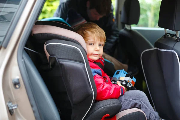 Πορτρέτο του όμορφου αγοριού που κάθεται στο κάθισμα του αυτοκινήτου. Ασφάλεια στη μεταφορά παιδιών. Ο πατέρας βάζει κλιπ σε έναν άλλο γιο στο βάθος. Χαριτωμένο υγιές παιδί αγόρι με παιχνίδι χαρούμενος για τις οικογενειακές διακοπές με το αυτοκίνητο — Φωτογραφία Αρχείου