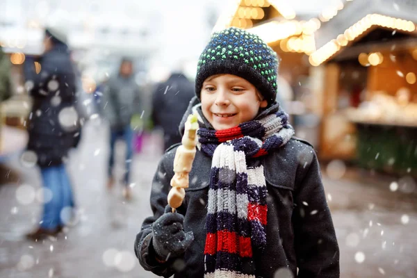 Little cute kid boy eating white chocolate covered fruits on skewer on traditional German Christmas market. Happy child on traditional family market in Germany during snowy day. — Stock Photo, Image