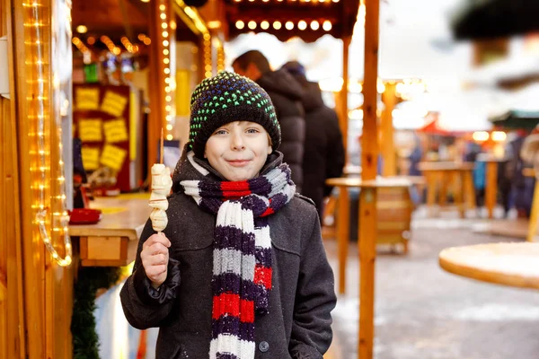 在传统的德国圣诞市场上 可爱的小男孩吃着白色的巧克力 上面涂满了扭曲的水果 雪天期间 德国传统家庭市场上的快乐孩子 — 图库照片
