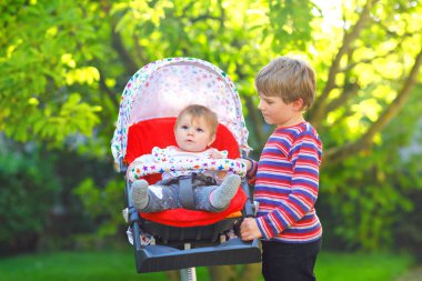 Küçük sarışın çocuk çocuk kardeşim ile oynamak. Mutlu kardeşler Bahçe. Çocuk arabası ya da bebek arabası oturan kız bebek. Kardeşim ve yaz gününde gidiş birlikte bir yürüyüşe şirin toddler açık havada