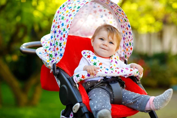 Leuke gezonde kleine mooie baby meisje zitten in de kinderwagen of kinderwagen en wachten op moeder. Gelukkig lachend kind met blauwe ogen. Met groene boomachtergrond. Kleine dochter gaat wandelen met familie — Stockfoto