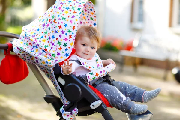 Linda niña hermosa y saludable sentada en el cochecito o cochecito y esperando a mamá. Feliz niño sonriente con ojos azules. Con fondo de árbol verde. Hija pequeña va a dar un paseo con la familia — Foto de Stock