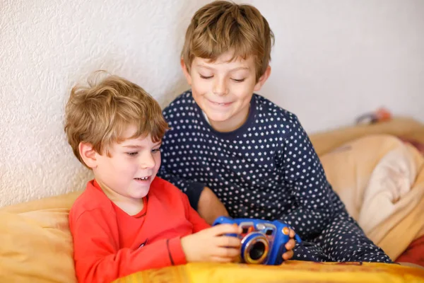 Два мальчика дошкольного или школьного возраста, братья и сестры веселятся после уроков, играя дома в видеоигры и фотографируясь на игрушечную камеру. Лучшие друзья, близнецы в пижаме или ночной одежде — стоковое фото