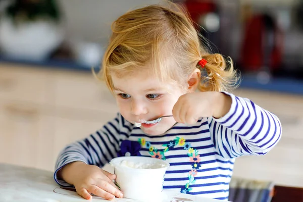 Entzückendes kleines Mädchen, das Hüttenkäse vom Löffel isst, gesunden Milchsnack. Nettes gesundes Kleinkind, Tochter mit Löffel sitzt im Hochstuhl und lernt allein in Küche oder Kinderzimmer zu essen — Stockfoto