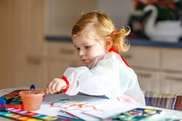 Симпатичная обожаемая девочка учится рисовать акварелью. Маленький ребенок рисует дома, используя красочные кисти. Здоровая счастливая дочь экспериментирует с цветами, водой дома или в детской — стоковое фото