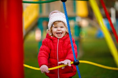 Küçük tatlı kız oyun alanında eğleniyor. Mutlu küçük çocuk tırmanışı, farklı ekipmanların üzerinde sallanmak ve kaymak. Renkli giysiler içinde soğuk bir günde. Çocuklar için aktif açık hava oyunu