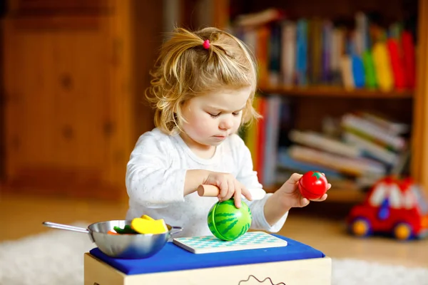 Schattig klein peutermeisje dat thuis speelt met eco houten speelgoed. Gelukkig gezond opgewonden kind snijden groenten en fruit met speelgoed mes. Baby meisje hebben plezier met rol keuken en koken spel. — Stockfoto