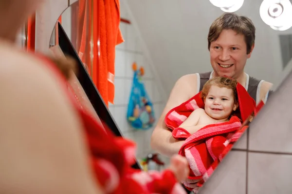 Pai segurando bonito menina criança embrulhado em toalha depois de tomar banho. Criança bebê saudável feliz com cabelos molhados olhando no espelho no banheiro e rindo. Relacionamento familiar de filha e pai — Fotografia de Stock