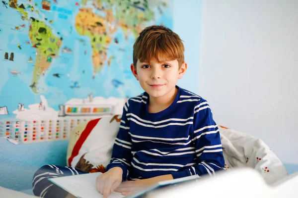 Lindo niño rubio en pijama leyendo un libro en su dormitorio. Emocionado niño sano leyendo alto, sentado en su cama. Colegial, familia, educación — Foto de Stock