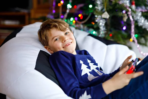 Niedlichen kleinen blonden Jungen spielen mit einem Videospiel auf Spielkonsole an Weihnachten mit geschmückten Baum auf dem Hintergrund. glücklich gesunde hild, die Spaß zu Hause hat. Beliebtes Weihnachtsgeschenk — Stockfoto