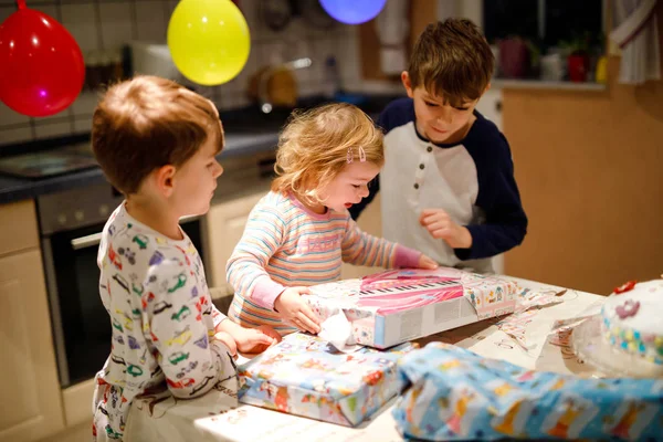 Очаровательная маленькая девочка, празднующая второй день рождения. Ребенок и два мальчика распаковывают подарки. Счастливые здоровые дети братья и сестры удивлены большими коробками и игрушками — стоковое фото