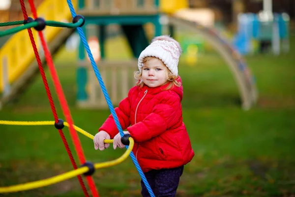 Nettes Kleinkind Mädchen hat Spaß auf dem Spielplatz. Glückliches gesundes kleines Kind klettern, schaukeln und rutschen auf verschiedenen Geräten. An kalten Tagen in bunten Klamotten. Aktives Outdoor-Spiel für Kinder — Stockfoto