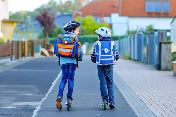 Två skolpojkar i skyddshjälm rider med skoter i staden med ryggsäck på solig dag. Glada barn i färgglada kläder cykling på väg till skolan. — Stockfoto