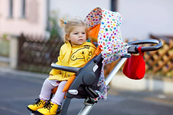 Retrato de una niñita mona sentada en un cochecito o cochecito y dando un paseo. Feliz bebé lindo que se divierte al aire libre. Hija sana. Tráfico callejero en la ciudad. — Foto de Stock