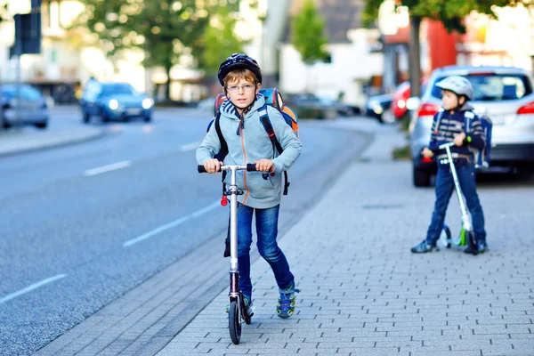 Zwei Schulkinder mit Schutzhelm fahren an einem sonnigen Tag mit dem Roller in der Stadt mit dem Rucksack. Fröhliche Kinder in bunten Kleidern radeln auf dem Schulweg. — Stockfoto