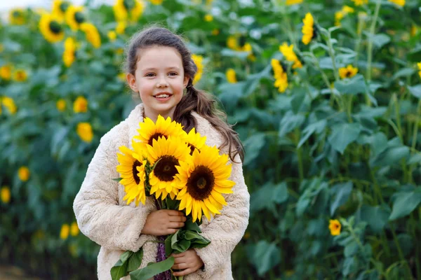 Roztomilá rozkošná školačka na slunečnicovém poli se žlutými květy. Krásné předškolní dítě s blonďatými vlasy. Šťastná zdravá dcerka, usmívající se a držící kytici — Stock fotografie