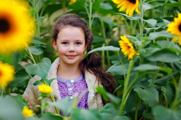 Aranyos iskolás lány a napraforgó pályán sárga virágokkal. Gyönyörű óvodás, szőke hajjal. Boldog egészséges kislányt, mosolyogva és csokorral a kezében — Stock Fotó
