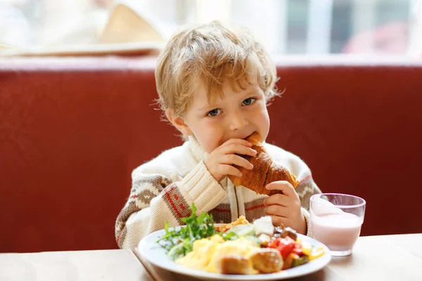 Rapaz saudável bonito comendo croissant e bebendo batido de morango no café. Criança feliz tomando café da manhã com os pais ou no berçário. Legumes, ovos como alimento saudável para crianças . — Fotografia de Stock