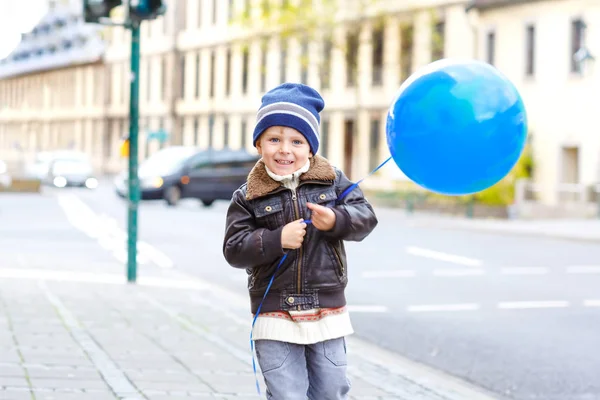 Menino bonito brincando com balão de ar azul ao ar livre na rua da cidade. Criança feliz correndo, andando, sorrindo e rindo. Criança saudável fora no dia frio em roupas quentes . — Fotografia de Stock