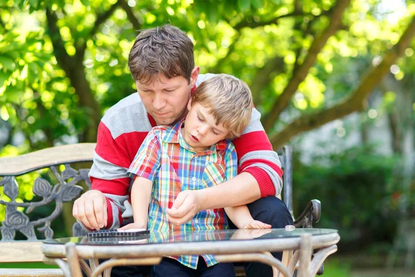 Il ragazzino e il suo giovane padre giocano insieme a dama. bambino e uomo trascorrere il tempo libero insieme. Famiglia si diverte nel giardino estivo all'aperto — Foto Stock