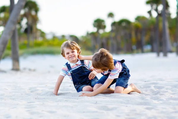 Dwoje małych chłopców bawiących się na tropikalnej plaży, szczęśliwi przyjaciele grający, koncepcja przyjaźni. Bracia rodzeństwo, bliźniaki walczące, biegające i skaczące w rodzinie z palmami na tle. — Zdjęcie stockowe
