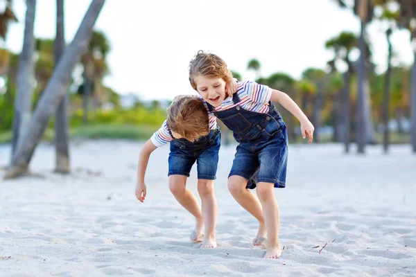 Dwoje małych chłopców bawiących się na tropikalnej plaży, szczęśliwi przyjaciele grający, koncepcja przyjaźni. Bracia rodzeństwo, bliźniaki walczące, biegające i skaczące w rodzinie z palmami na tle. — Zdjęcie stockowe