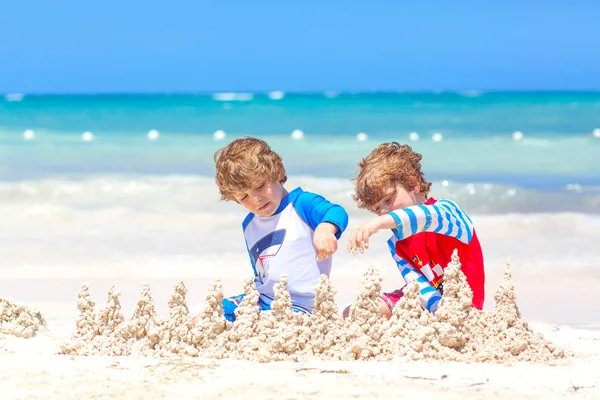Два маленьких мальчика веселятся, строя песчаный замок на тропическом пляже на острове. Здоровые дети играют вместе в отпуске. Близнецы, счастливые братья смеются и улыбаются — стоковое фото