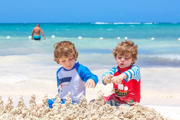 İki küçük çocuk adada tropikal plajda kumdan kale inşa ederken eğleniyor. Tatillerinde birlikte oynayan sağlıklı çocuklar. İkizler, Mutlu kardeşler gülüyor ve gülümsüyor — Stok fotoğraf