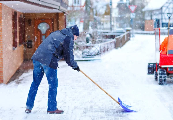 Kar kürekli bir adam kar yağışı sırasında kaldırımları temizler. Avrupa 'da kış mevsimi. Sıcak kış kıyafetleri giymiş genç bir adam. Almanya 'da kar ve hava kaosu. Kar fırtınası ve yoğun kar. Schneechaos — Stok fotoğraf