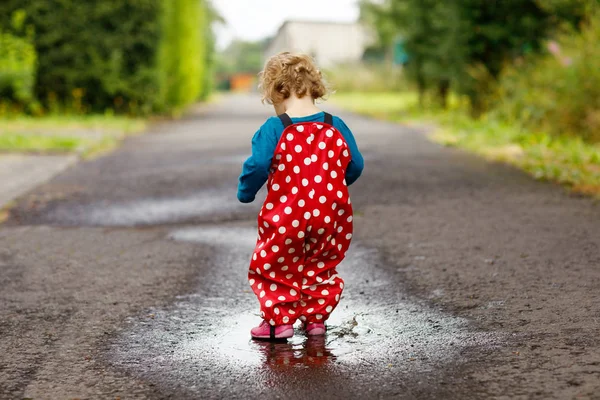 Närbild av lilla småbarn flicka klädd regn stövlar och byxor och promenader under snöblandat regn, regn på kall dag. Baby barn i färgglada Mode casual kläder hoppa i en vattenpöl — Stockfoto