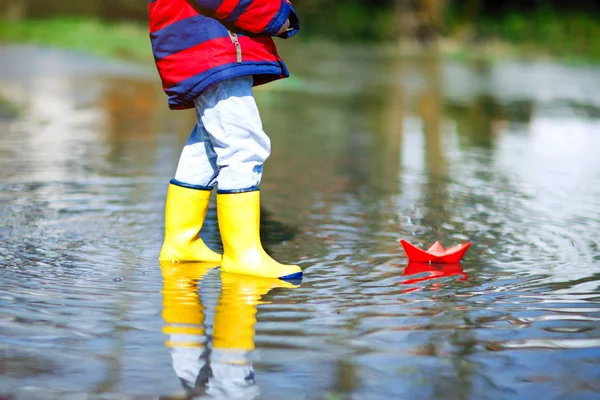 春天或秋天，穿着黄色雨靴的快乐小男孩在大水坑边玩纸船 — 图库照片