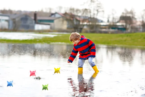 Happy little kid boy in yellow rain boots playing with paper ship boat by huge puddle on spring or autumn day — Stock Photo, Image