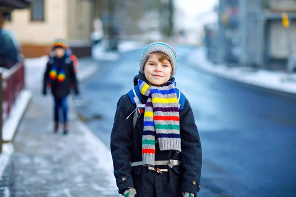 Dva malí kluci ze základní třídy chodili během sněžení do školy. Šťastné děti se baví a hrají si s prvním sněhem. Sourozenci přátelé s batohem v barevných zimních šatech. — Stock fotografie