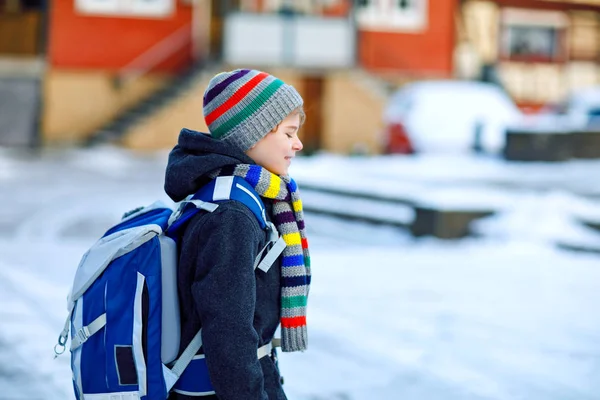 Malý školáček základní třídy, který chodí do školy během sněžení. Šťastné dítě baví a hrát si s prvním sněhem. Student s batohem nebo brašnou v barevných zimních šatech. — Stock fotografie